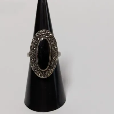 Sale Photo Thumbnail #791: Vintage sterling silver ring featuring a central oval black onyx stone surrounded by a marcasite halo, displayed on a ring stand.