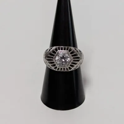 Sale Photo Thumbnail #808: Art Deco style filigree ring featuring a central round clear stone set within an intricate hexagonal geometric framework, displayed on a black ring cone.
