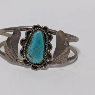 Sale Photo Thumbnail #821: Vintage Native American sterling silver cuff bracelet featuring a central turquoise cabochon flanked by hand-engraved leaf accents.