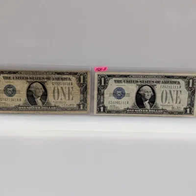 Sale Photo Thumbnail #839: Two Series 1928 one-dollar silver certificates, featuring blue treasury seals and housed in protective plastic sleeves.