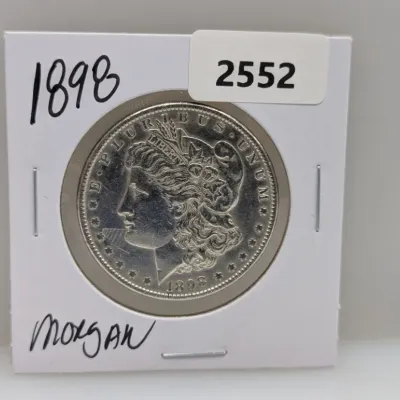 Sale Photo Thumbnail #877: 1898 Morgan silver dollar coin housed in a protective cardboard flip.