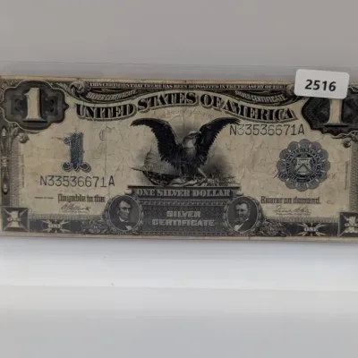 Sale Photo Thumbnail #841: Series 1899 $1 'Black Eagle' Silver Certificate housed in a protective plastic sleeve, featuring portraits of Abraham Lincoln and Ulysses S. Grant.
