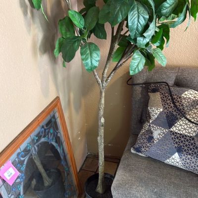 Sale Photo Thumbnail #19: Tall faux potted ficus tree set in a black nursery pot, shown alongside a framed leaded glass artwork and a tufted grey accent chair.