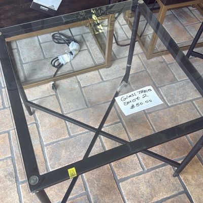 Sale Photo Thumbnail #30: Contemporary glass-top end table with a black metal frame and X-brace support, shown with dark wood and brass accent furniture in the background.