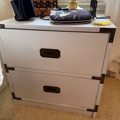 Sale Photo Thumbnail #27: White campaign-style two-drawer file cabinet with dark metal corner brackets and recessed handles, topped with assorted home goods and electronics.