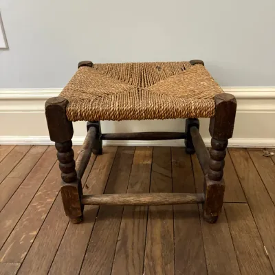 Sale Photo Thumbnail #112: Rustic rustic footstool featuring turned wooden legs and a woven rush seat, showing minor wear consistent with age.