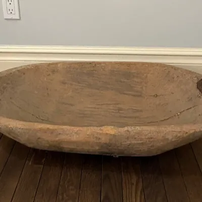 Large antique hand-carved wooden dough trough resting on a hardwood floor, featuring a rustic weathered patina and old metal staple repairs.