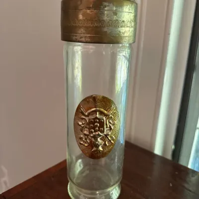 Vintage glass decanter or jar featuring an embossed brass crest emblem and a decorative brass cap.