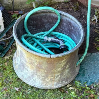 Vintage tarnished metal planter tub repurposed as a garden hose holder, featuring a coiled green hose and spray nozzle inside.