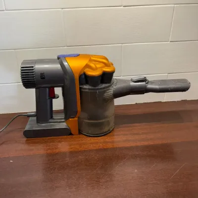 Sale Photo Thumbnail #149: Dyson DC34 handheld cordless vacuum cleaner with charging cable attached, sitting on a wooden countertop.