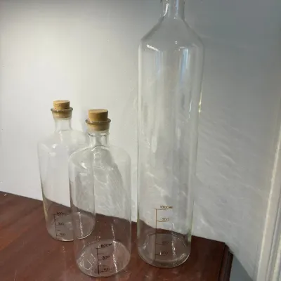 Sale Photo Thumbnail #111: Set of three laboratory-style clear glass apothecary bottles with cork stoppers and metric volume measurement markings.