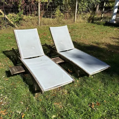 Pair of outdoor teak chaise lounge chairs with light grey mesh seating and pull-out side trays, showing weathered patina.