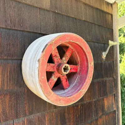 Vintage heavy-duty industrial pulley wheel with chipped red paint, mounted on a wood-shingled exterior wall.