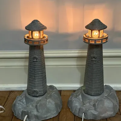 Sale Photo Thumbnail #155: Pair of vintage lighthouse novelty lamps with painted stone-effect finish and glowing top lights, set on rocky bases.