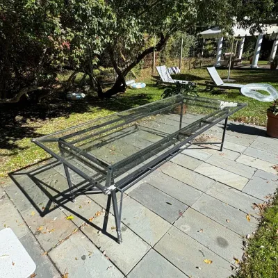 Large rectangular outdoor dining table featuring a dark metal frame and a multi-pane glass tabletop, positioned on a flagstone patio.