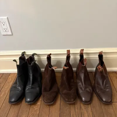 Three pairs of R.M. Williams Chelsea boots lined up on a hardwood floor, including black leather, brown suede, and dark brown leather...