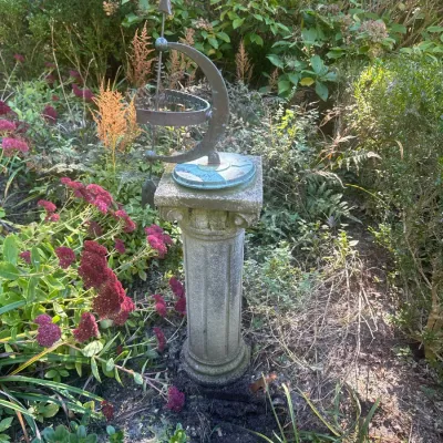Vintage brass armillary sphere sundial with gnomon arrow mounted on a weathered stone pedestal column in a garden setting.