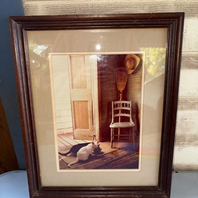 Sale Photo Thumbnail #392: Framed photographic art print featuring a porch scene with two cats, a chair, straw hat, and broom. Housed in a dark wooden frame with double matting under glass. Frame shows light age-appropriate wear and minor scuffing around the edges.
15.5X12.5in