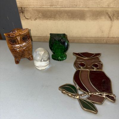 Sale Photo Thumbnail #19: Collection of four owl-themed decorative items, including a carved wooden owl, a green glass owl figurine, a clear glass or crystal owl paperweight, and a stained glass owl sun catcher on a branch. All items appear to be in good condition with no visible 
