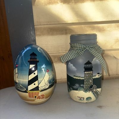 Sale Photo Thumbnail #344: A decorative two-piece nautical lot featuring hand-painted lighthouse scenes. Includes one large glossy painted wooden egg featuring a coastal lighthouse with sailboats, and one painted glass mason jar decor piece with a lighthouse motif tied with a green