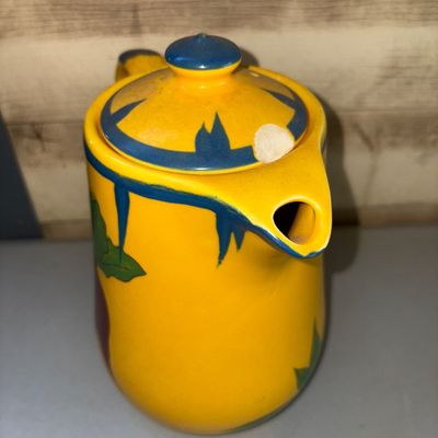 Sale Photo Thumbnail #218: Vibrant yellow ceramic pitcher or teapot featuring colorful hand-painted folk art illustrations including a figure with a pipe under a palm tree and a smiling sun motif. Bright blue glaze accents trim the spout, rim, lid, and handle. Note: There is a nota