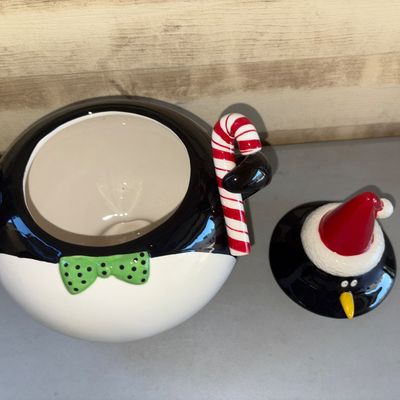 Sale Photo Thumbnail #200: Ceramic festive penguin-shaped cookie jar featuring a Santa hat lid, green polka dot bow tie, and holding a candy cane. The piece has a glossy painted finish. In good pre-owned condition with no visible chips or cracks.
11in tall