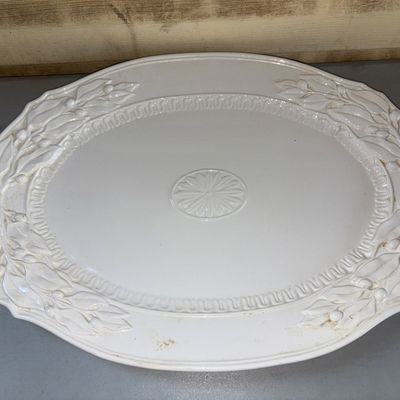 Sale Photo Thumbnail #381: Large oval ceramic or porcelain serving platter in an off-white/cream glaze. Features embossed leaf and berry sprigs along the scalloped rim, an inner dentil/guilloche border, and a central floral rosette medallion. Shows light surface wear and minor disc