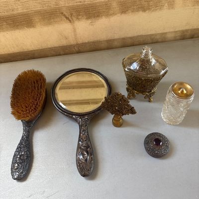 Sale Photo Thumbnail #10: A 4-piece vintage vanity collection featuring an ornate embossed metal hand mirror with a floral design on the reverse, a matching natural bristle hairbrush, an ornate brass or gold-tone filigree perfume bottle with a decorative stopper, and a cut glass v