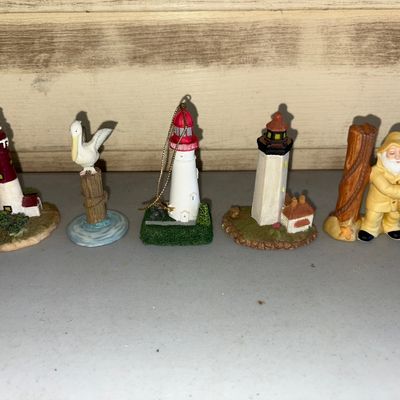 Sale Photo Thumbnail #138: Collection of five miniature nautical-themed resin figurines. Includes three lighthouses (one with a hanging cord/ornament loop), a pelican perched on a wooden piling, and a fisherman in yellow rain gear. All pieces appear to be in good pre-owned conditio