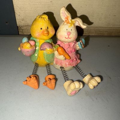 Sale Photo Thumbnail #273: Pair of painted resin or composite Easter shelf sitter figurines featuring a yellow chick holding a basket of eggs and a pink-dressed bunny holding a carrot. Both feature flexible metal spring legs with feet designed to dangle over an edge. Figures show l