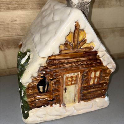 Sale Photo Thumbnail #47: 8X9in.
Glazed ceramic cookie jar shaped like a rustic winter log cabin with a snow-covered roof, chimney, lantern, and evergreen tree details. Features a removable roof lid. In good condition with no visible cracks or major chips.