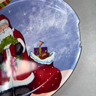 Sale Photo Thumbnail #320: Set of two festive plastic/melamine Christmas serving trays featuring Santa Claus designs. One oval tray with a blue snowy background shows noticeable damage and chipping along the right edge. The second green scalloped tray features Santa holding a prese