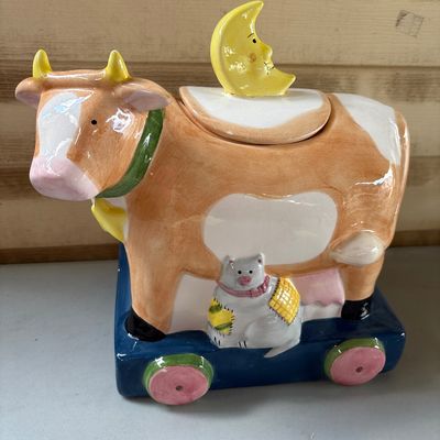 Sale Photo Thumbnail #84: Charming hand-painted ceramic cookie jar featuring a cow on a pull-toy style base with a cat, milk pail detail, and a crescent moon lid handle inspired by the nursery rhyme. Shows minor paint wear and small chips on the moon handle tip and horn.
10X8X10in