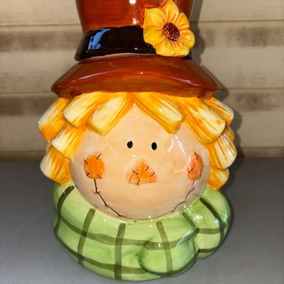 Sale Photo Thumbnail #159: A glazed ceramic jar featuring a scarecrow wearing a green plaid scarf and a brown hat with a sunflower detail. The hat serves as a removable lid. Glazed ceramic finish in bright autumn colors. Good condition with no visible chips or cracks.
10in tall