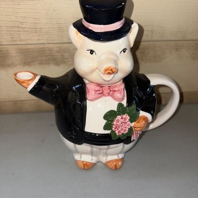 Sale Photo Thumbnail #54: Novelty ceramic teapot shaped like a dapper pig dressed in a black top hat, tuxedo coat, pink bow tie, and holding a small bouquet of flowers. Features a spout formed by one arm and a rear handle. Appears to be in good pre-owned condition with no visible 