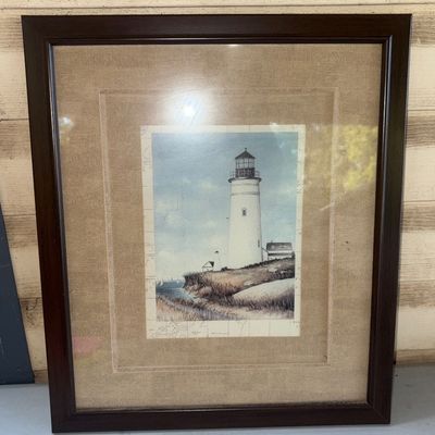 Sale Photo Thumbnail #359: Framed artwork depicting a lighthouse on a coastal cliff set over a subtle map background. Features double matting with a textured woven outer mat and a dark brown wood frame. Protected under glass. Appears to be in good condition overall with minor wear 