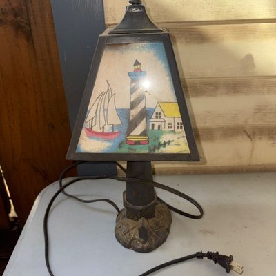Sale Photo Thumbnail #353: Small vintage nautical-themed electric table lamp featuring a cast metal lighthouse-style base with a simulated stone ground and a metal-framed trapezoidal shade. The shade panels depict a colorful coastal scene with a lighthouse, sailboat, and house. Cor
