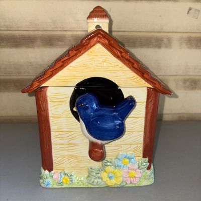 Sale Photo Thumbnail #178: Glazed ceramic cookie jar or canister shaped like a birdhouse, featuring a bluebird perched at the entrance and colorful floral details along the base. Removable roof lid. Appears to be in good pre-owned condition with no visible cracks or chips.
10in tal