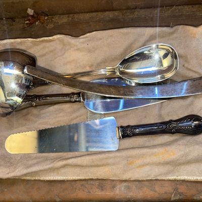Sale Photo Thumbnail #274: Collection of silver-tone/silverplate serving pieces including a soup ladle, large serving spoon, cake/pie servers (one with a serrated edge), and a curved serving piece. Includes a fabric storage pouch. Items show varying degrees of surface wear, scuffin