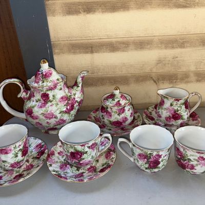 Sale Photo Thumbnail #357: A porcelain tea set decorated with an all-over pink rose pattern and gold accents. The set includes a covered teapot, a covered sugar bowl with saucer, a cream pitcher with saucer, and four teacups (two with matching saucers). Appears to be in good condit