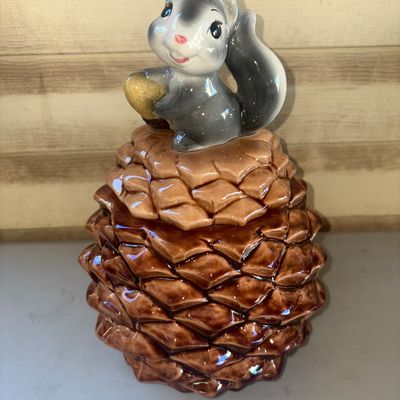 Sale Photo Thumbnail #260: A charming vintage ceramic cookie jar designed as a brown pinecone, featuring a removable lid topped with a grey squirrel figurine holding an acorn. High-gloss glazed finish throughout. Appears to be in good vintage condition with no major chips or cracks