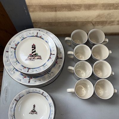 Sale Photo Thumbnail #307: A comprehensive ceramic dinnerware collection featuring a coastal lighthouse pattern with sailboat borders. The set includes large dinner plates, salad/salad plates, soup/cereal bowls, and matching coffee mugs. Items appear to be in good used condition wi