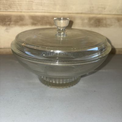 Sale Photo Thumbnail #143: Clear glass covered candy or trinket dish with a pedestal base and ribbed texture detailing around the lower bowl. Features a fitted lid with a raised central knob. Appears to be in good pre-owned condition with minor surface wear consistent with age.
5in