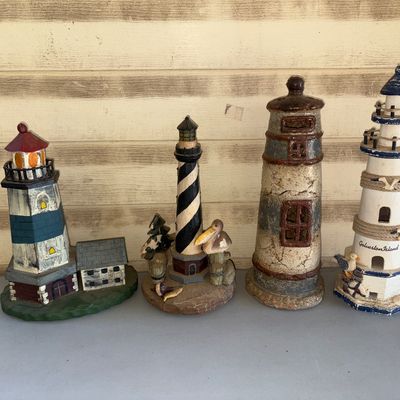 Sale Photo Thumbnail #150: A collection of four decorative lighthouse figurines in various styles and materials, including resin and wood. Features coastal details such as seagulls, pelicans, rope trim, and one labeled 'Galveston Island'. Items are in good pre-owned condition with 