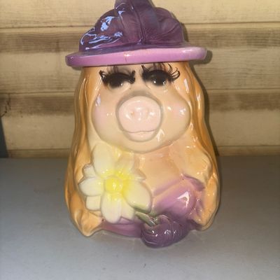 Sale Photo Thumbnail #247: Vintage ceramic cookie jar featuring Miss Piggy wearing a purple hat and holding a white and yellow daisy. Includes removable purple hat lid. Overall good condition with glossy finish; minor wear and light paint imperfections consistent with age and use.
