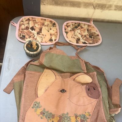 Sale Photo Thumbnail #292: A collection of pig-themed kitchen items featuring a fabric apron with a large pig applique front pocket, two matching quilted potholders with printed pig motifs, and a small decorative pig-themed mechanical kitchen timer. Items show minor signs of wear c