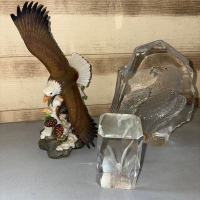 Sale Photo Thumbnail #38: A collection of three eagle-themed decorative items. Includes a hand-painted resin bald eagle figurine perched on a branch base with pinecones, a large clear glass sculpture with an etched/molded eagle profile, and a clear glass block featuring a 3D laser