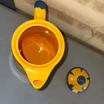 Sale Photo Thumbnail #219: Vibrant yellow ceramic pitcher or teapot featuring colorful hand-painted folk art illustrations including a figure with a pipe under a palm tree and a smiling sun motif. Bright blue glaze accents trim the spout, rim, lid, and handle. Note: There is a nota
