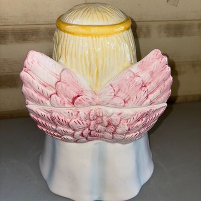 Sale Photo Thumbnail #165: Charming ceramic praying angel cookie jar with lid. Features pastel blue and white robes, blonde hair with a yellow halo, and pink feathered wings. Glazed finish. Shows light wear consistent with age, including subtle hairline cracking near the top rim of