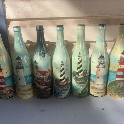 Sale Photo Thumbnail #342: Collection of seven decorative glass wine bottles, each hand-painted with detailed lighthouse and coastal scenes. Features various lighthouse designs including spiral stripe and red-and-white striped patterns with ocean backdrops. Shows minor paint wear a